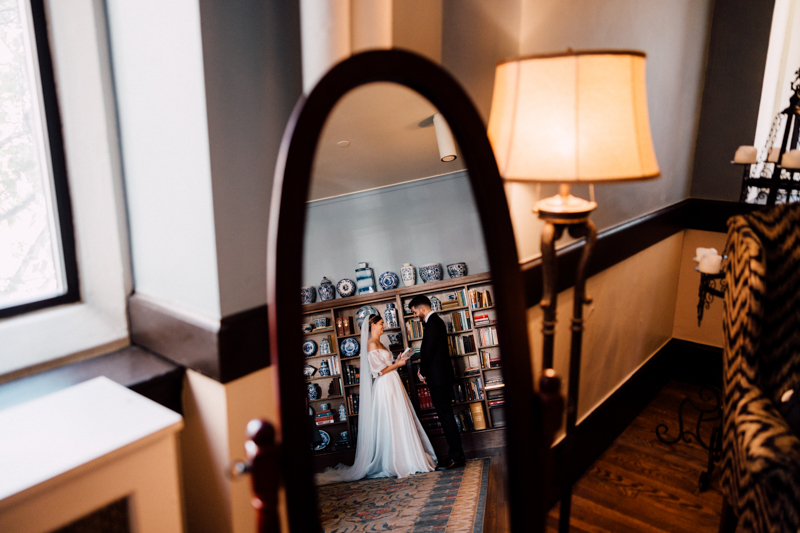 A bride and groom stand close together in front of a bookshelf at the Pavilion at Grace Wedding, their image reflected in a tall mirror. The cozy room has warm lighting, a lamp, and decorative shelves in the background.