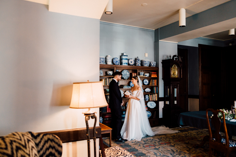 A bride and groom stand together holding hands in a warmly lit room with blue walls, a lamp, and patterned rug, reminiscent of the elegance found at a Pavilion at Grace Wedding, with shelves of decorative blue and white vases behind them.