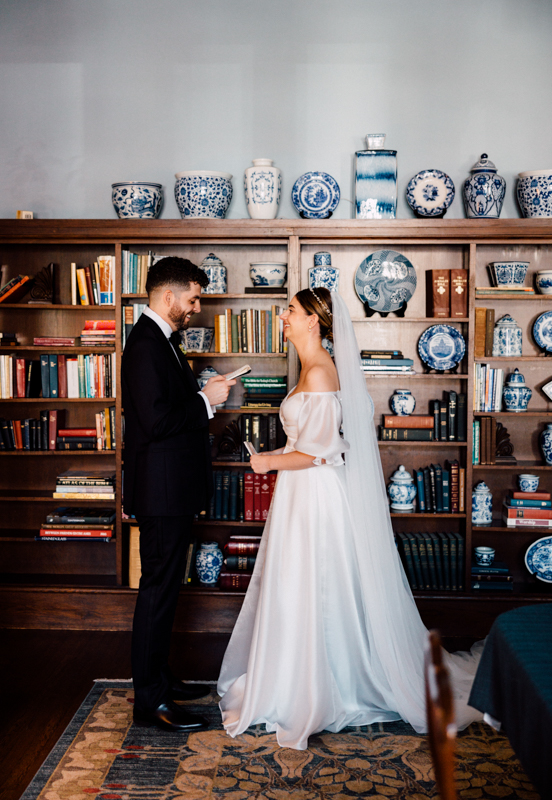 At the Pavilion at Grace Wedding, a groom in a black suit reads vows to a smiling bride in an off-shoulder white gown and veil, standing before a bookshelf adorned with blue and white vases and books.