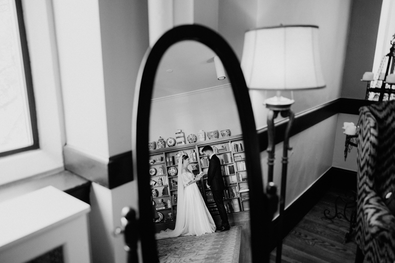A black and white photo shows a bride and groom holding hands, reflected in a standing mirror at the Pavilion at Grace Wedding. Behind them, a bookshelf filled with various items creates an intimate and romantic scene.