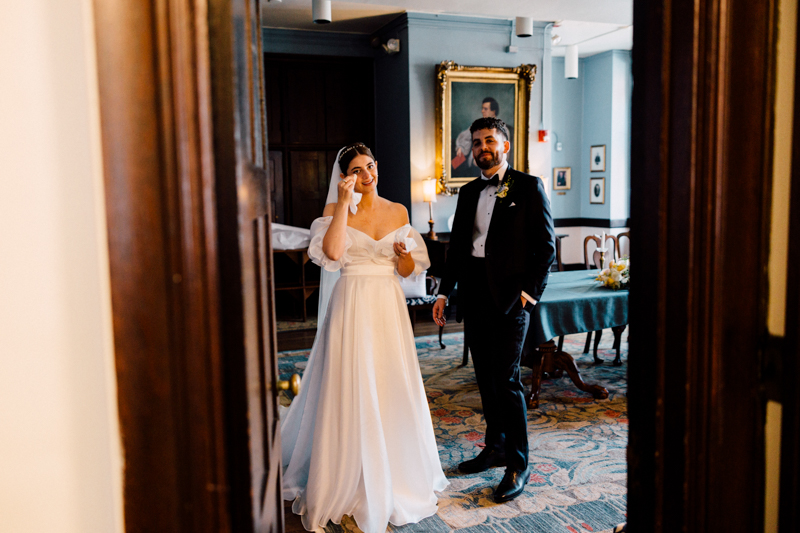 A bride in a white gown wipes a tear from her face while standing next to a groom in a black tuxedo at the Pavilion at Grace Wedding, surrounded by warm light, classic decor, and elegant paintings on the walls.