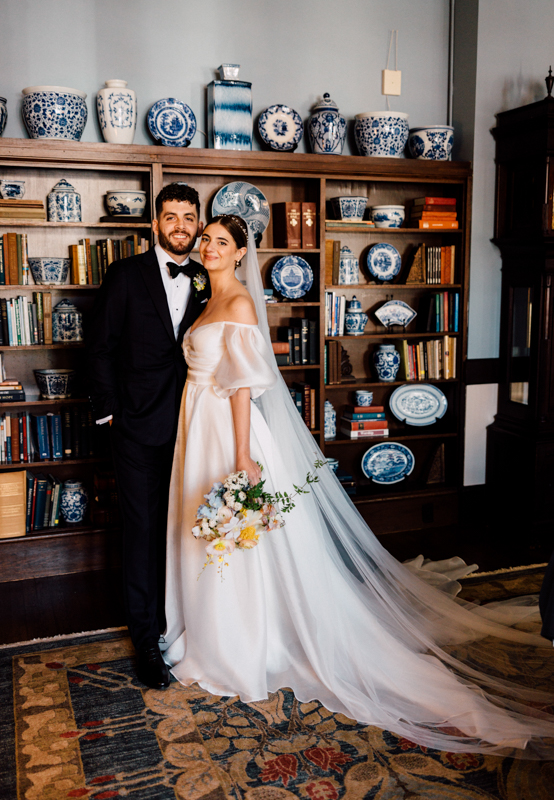 A bride in an off-shoulder white gown and veil stands smiling beside a groom in a black tuxedo at the Pavilion at Grace Wedding, posing before a wooden bookshelf filled with blue and white china and books. The bride holds a bouquet of flowers.