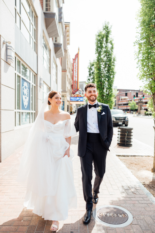 A bride in a white dress and veil walks beside a groom in a black tuxedo. Smiling, they stroll on a city sidewalk in daylight, with buildings and a tree behind them—capturing the joy of their Pavilion at Grace Wedding.