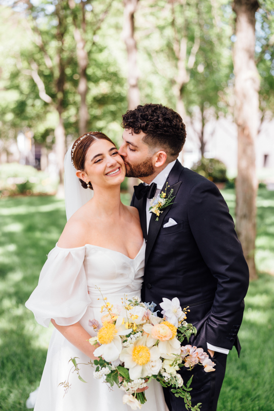 A groom in a black tuxedo kisses his smiling bride in an off-the-shoulder white gown as she holds a bouquet of yellow and white flowers, captured at the sunlit Pavilion at Grace Wedding amidst lush greenery.