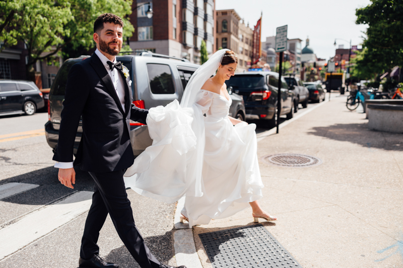 A bride in a white dress and veil walks with her groom in a black tuxedo on a city sidewalk, smiling and holding her dress—moments before their Pavilion at Grace Wedding—with parked cars and buildings in the background.