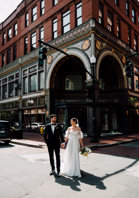 A bride and groom in wedding attire hold hands and walk across a city street under a green light, with the historic red-brick Pavilion at Grace Wedding venue and its large arched entryway in the background.