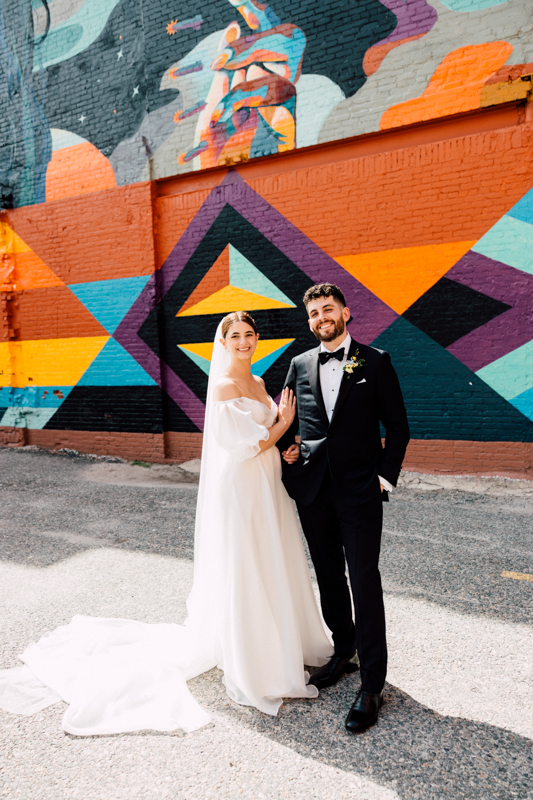 A bride in a white gown and veil stands beside a groom in a black tuxedo, both smiling, at their Pavilion at Grace Wedding, in front of a colorful geometric mural painted on a brick wall.