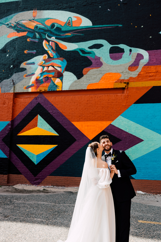 A bride and groom smile and embrace in front of a colorful mural at their Pavilion at Grace Wedding, featuring geometric shapes, an astronaut’s hand, and a plane. The bride wears a white dress and veil; the groom wears a black tuxedo.