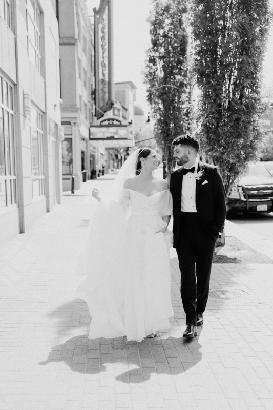 A bride in a flowing white gown and veil walks arm in arm with her groom in a tuxedo on a city sidewalk near the Pavilion at Grace Wedding, smiling at each other. The scene is captured in timeless black and white.