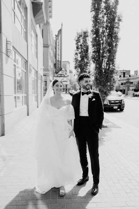 A bride in a white wedding dress and veil stands smiling beside a groom in a black tuxedo as they walk hand in hand outside the Pavilion at Grace Wedding, enjoying a sunny day on the city sidewalk.