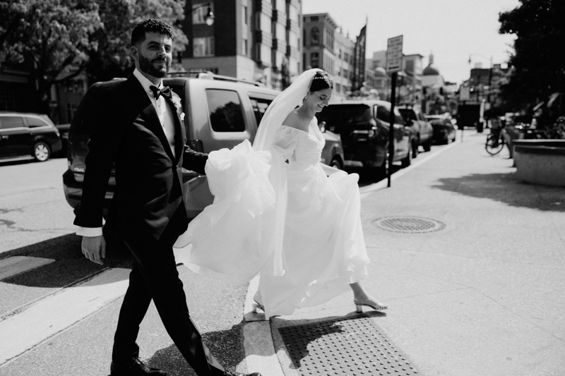A bride in a flowing white dress and veil walks beside her groom on a city street, holding up her gown as they cross. The black-and-white photo captures an urban Pavilion at Grace Wedding moment.