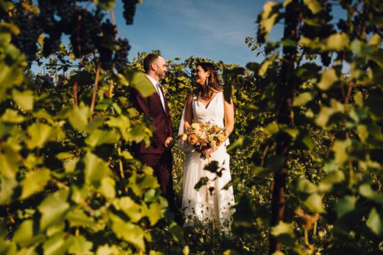 A bride and groom stand together in a vineyard at their Shelburne Vineyard Wedding, surrounded by lush green grapevines. The bride holds a bouquet of flowers as they smile at each other under a clear blue sky.