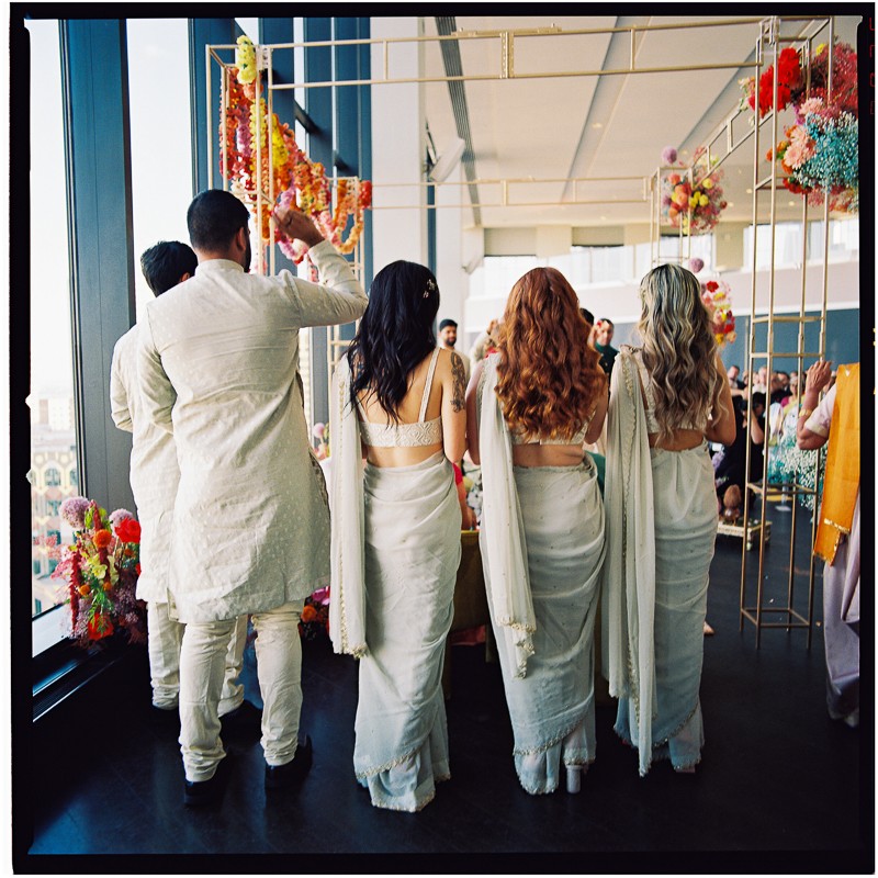 Four people dressed in white traditional South Asian attire stand with their backs to the camera at a vibrant State Room Boston wedding, captured on 120mm film, colorful flowers and guests completing the elegant indoor ceremony scene.