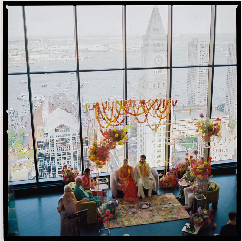 A couple in traditional clothing sits under a colorful floral mandap at their State Room Boston wedding, captured on 120mm film, with cityscape and waterfront views shining through the large windows.