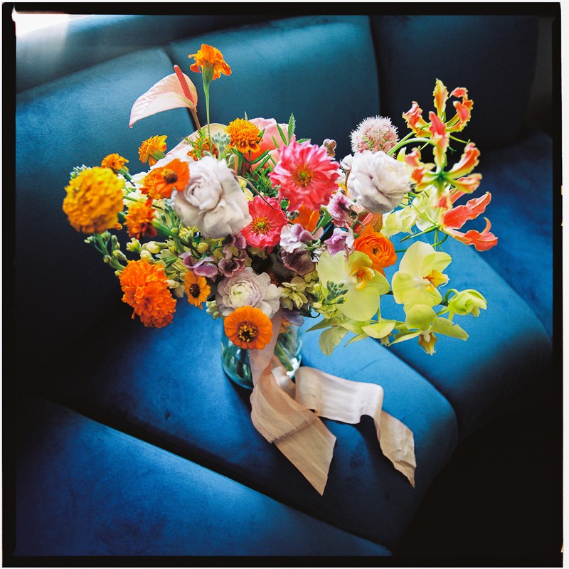 A vibrant bouquet of assorted flowers in a glass vase on a blue velvet couch with natural light highlighting the arrangement.