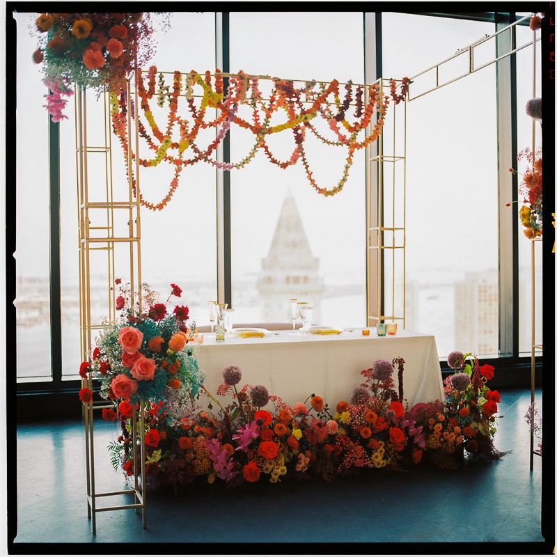 An elaborately decorated event space with a table adorned in a vibrant floral arrangement, set against floor-to-ceiling windows overlooking a cityscape. A decorative floral garland hangs above, adding a festive atmosphere to the scene.