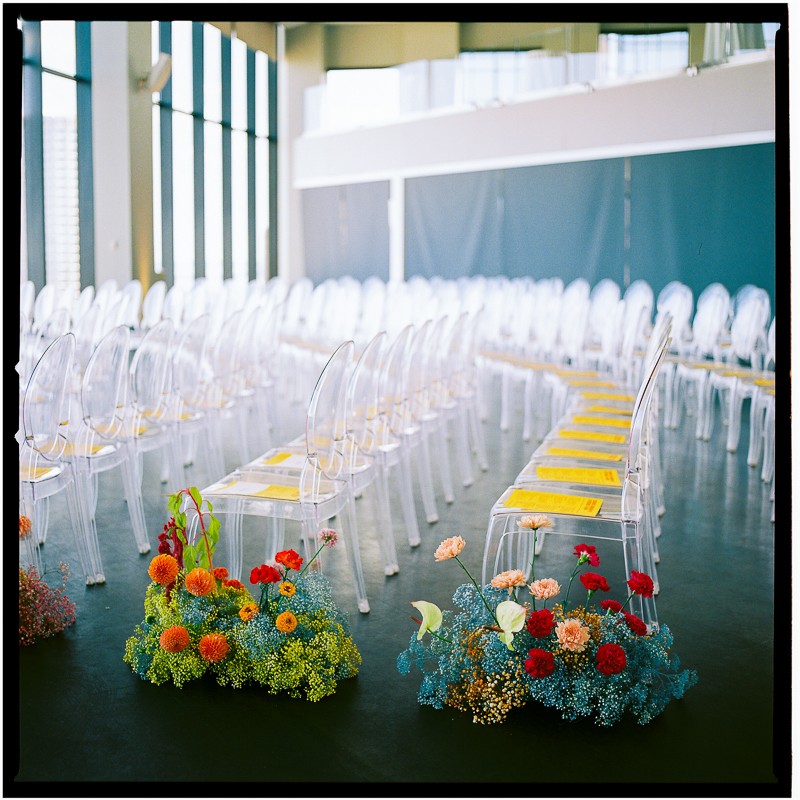 An event space with rows of clear acrylic chairs set up for a ceremony, with vibrant floral arrangements decorating the aisle.
