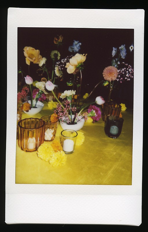 A Polaroid photo depicting a variety of colorful flowers arranged in multiple vases on a table covered with a yellow cloth, accompanied by candles and decorative glasses.