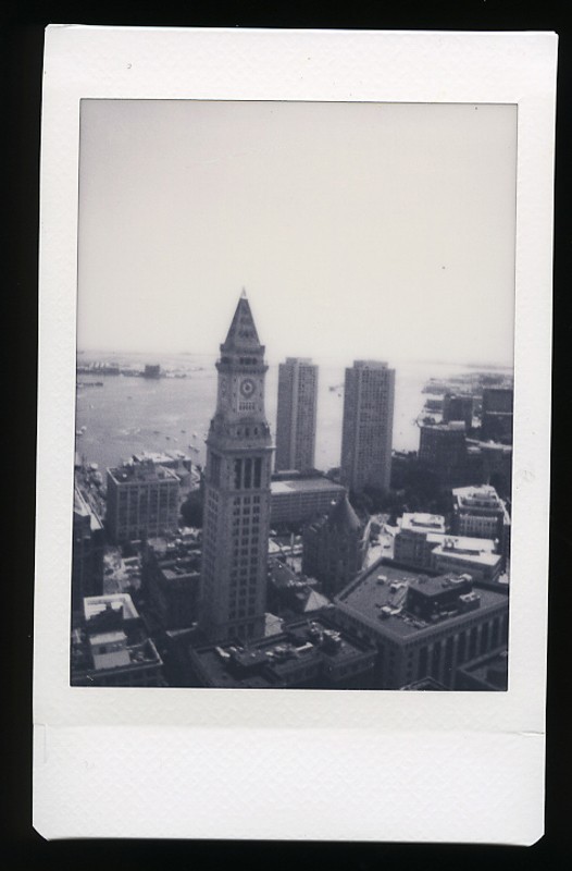 A Polaroid photo depicting an aerial view of a historic clock tower in an urban setting with buildings surrounding it and a body of water in the background. The image has a monochromatic, vintage appearance.
