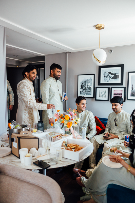 A group of people in traditional South Asian attire gathered in a cozy living room with an array of food on the table, engaging in conversation and enjoying each other's company.