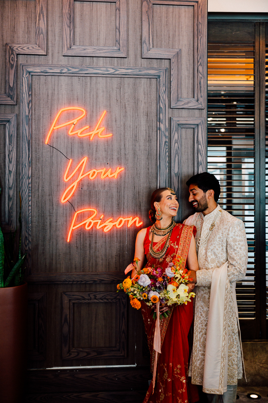 A happy couple in traditional South Asian attire standing in front of a wooden background with a neon sign that reads "Pick Your Poison." The woman is holding a vibrant bouquet of flowers.