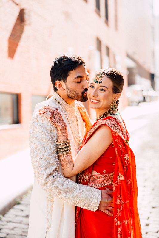A couple in traditional Indian attire embraces outdoors. The man kisses the woman’s temple as she smiles, wearing a red sari with gold details and green jewelry. Sunlight brightens the scene, captured by a State Room Boston Wedding 35mm film photographer.