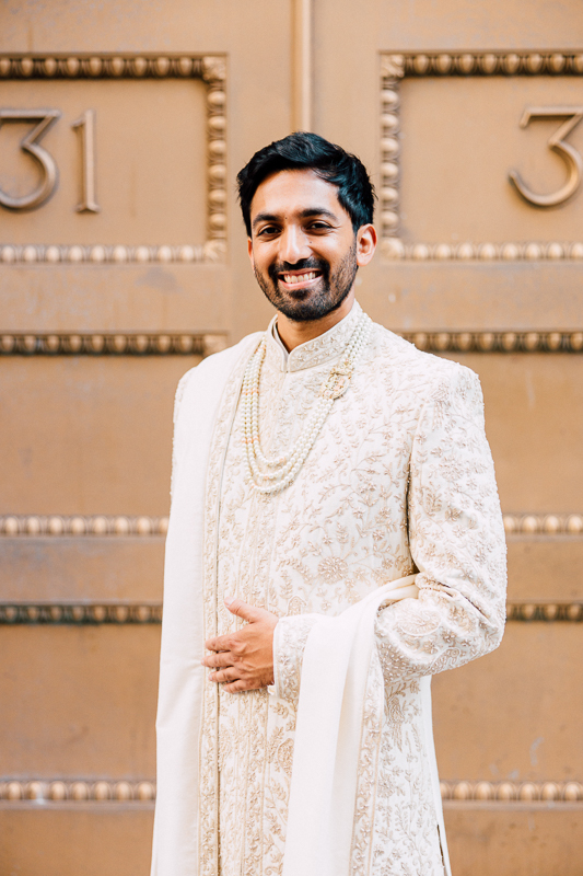 A smiling man wearing a traditional embroidered cream sherwani with a string of pearls, standing in front of a building with the number 31.
