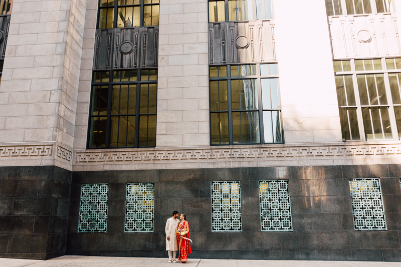 A couple stands in front of a large, decorative building with geometric patterns on the facade and ornate window grills. The man is dressed in a white outfit, while the woman wears a red and gold sari. They appear to be engaged in conversation.