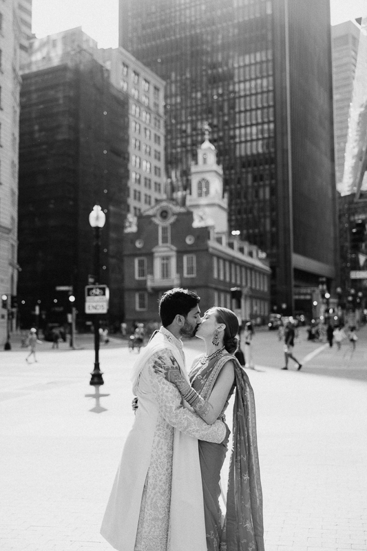 A couple dressed in traditional South Asian wedding attire sharing an intimate moment in a sunlit urban setting with buildings and a historic structure in the background. The image is in black and white.