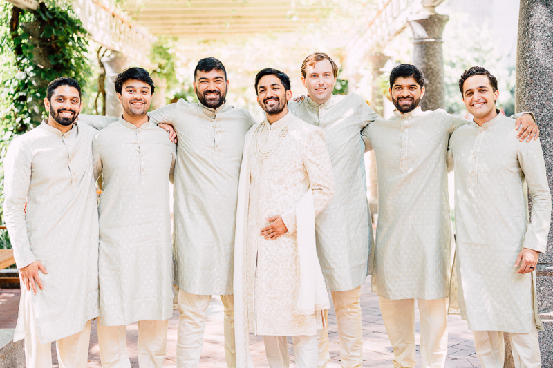 A group of seven smiling men, dressed in traditional cream-colored Indian attire, standing arm in arm outdoors with trees and a vine-covered trellis in the background.
