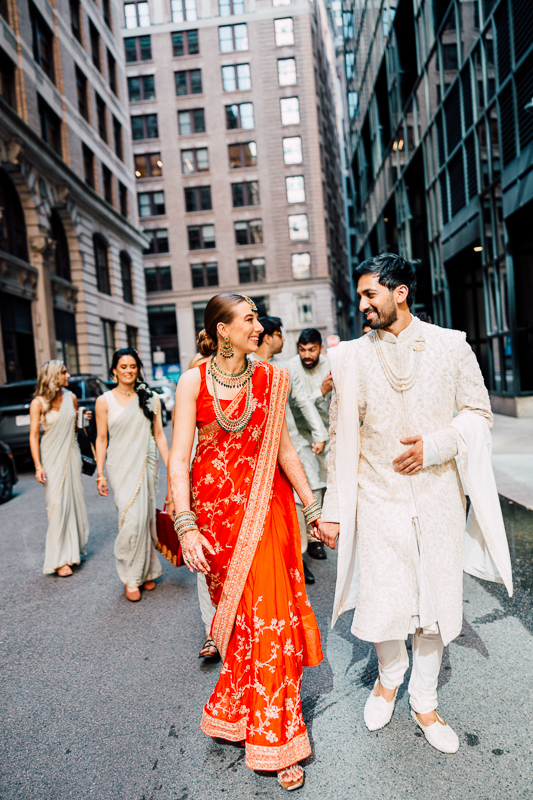 A joyful couple in traditional Indian wedding attire walks hand in hand down a city street with a group of people in the background.