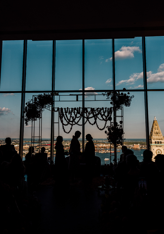 A silhouetted wedding ceremony unfolds indoors at the State Room Boston, with city and river views; floral decorations and a canopy frame the couple and guests under a blue sky, all beautifully captured by a wedding film photographer.