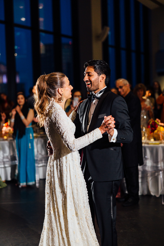 A bride in a white dress and a groom in a black tuxedo smile and dance together at their State Room Boston wedding, captured beautifully by a film photographer, with guests and decorated tables in the background.