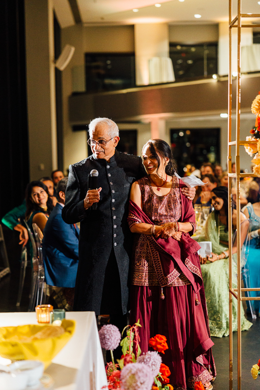 A smiling older couple standing together at a social event, the man holding a microphone and the woman holding a piece of paper, both dressed in traditional Indian attire. There is a seated audience in the background and the atmosphere appears festive with floral decorations.