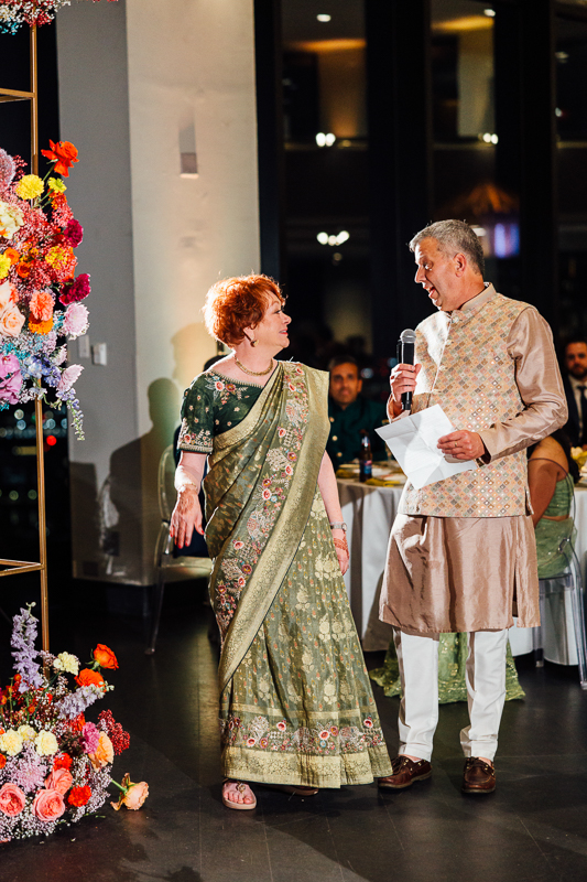 A man and a woman dressed in traditional Indian attire are conversing at a social event, with the woman looking animatedly at the man who is holding a microphone and some papers. They are standing near a colorful floral arrangement.