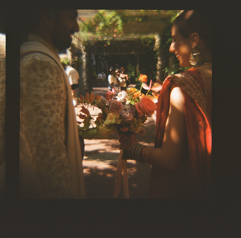 Alt text: A man and woman in traditional Indian attire partially facing each other with the woman holding a vibrant bouquet of flowers, all bathed in warm, natural light with a dreamy, vignette border.