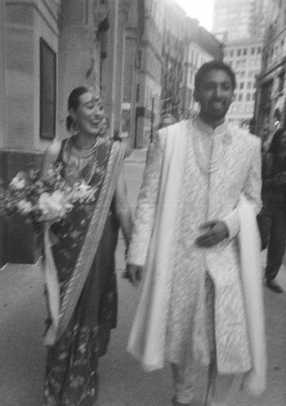A black and white photo by a State Room Boston Wedding 35mm film photographer captures a couple in traditional South Asian wedding attire, walking outdoors hand in hand and smiling. The woman holds a bouquet, with tall buildings and blurred figures in the background.