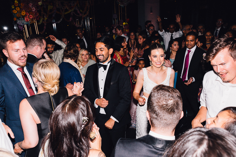 A joyful bride and groom holding hands as they walk through a crowd of guests at a wedding reception, with some guests dancing and others looking on, all under bright festive lights.