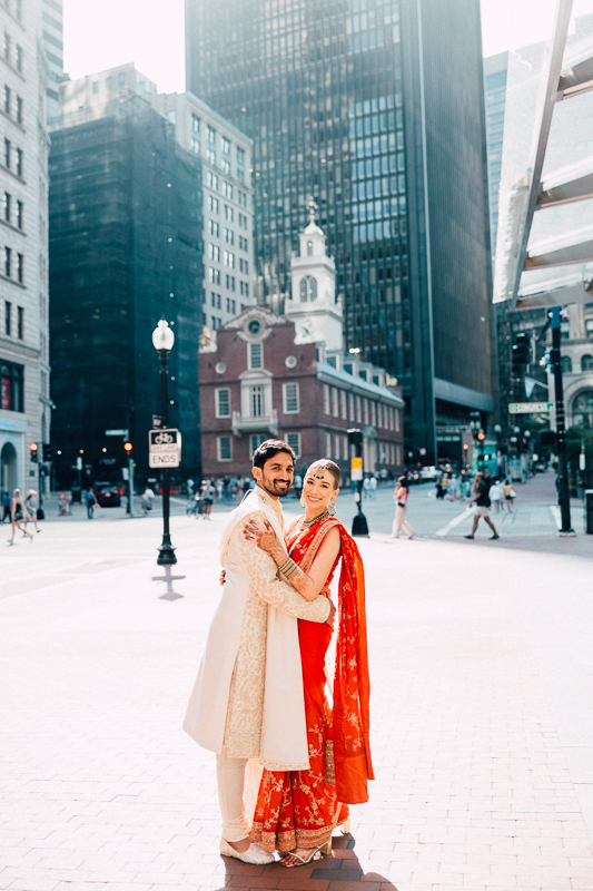 A couple dressed in traditional South Asian wedding attire is embracing and smiling in an city square with buildings and a few pedestrians in the background.