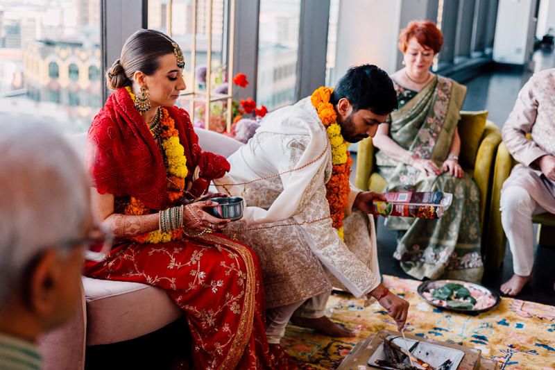 A traditional South Asian wedding ceremony with a bride in a red sari and groom in a cream sherwani, both adorned with marigold garlands, performing a ritual with a fire in front of seated guests.