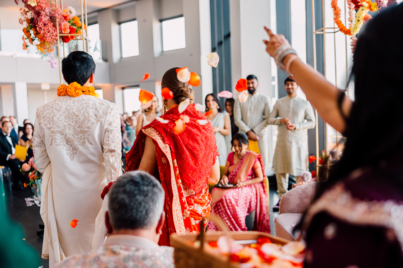 A bride and groom in traditional South Asian wedding attire stand with their backs to the camera. Petals are tossed in the air around them by guests, creating a joyous atmosphere. The couple is flanked by seated and standing attendees in colorful clothing, in a bright venue decorated with floral arrangements.