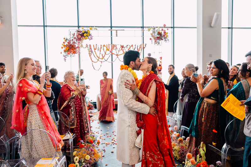 A newlywed couple in traditional Indian attire shares a kiss at the end of their State Room Boston wedding aisle, surrounded by smiling guests applauding and capturing the moment, with colorful flowers and garlands decorating the venue.