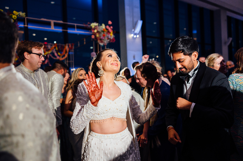 A woman in a white embellished outfit, with henna on her hands, smiles and lifts her hands while dancing at a lively indoor State Room Boston wedding, captured by a film photographer among guests in formal and traditional attire.