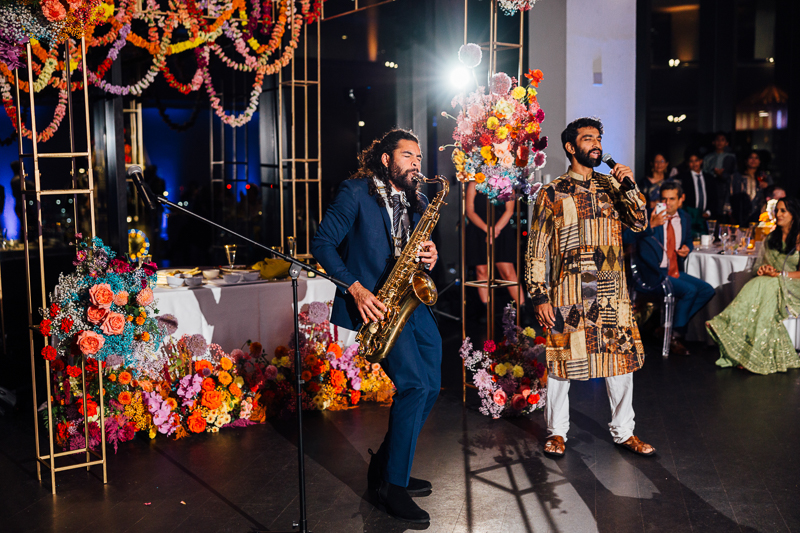 Two men perform at an event in the State Room Boston; one plays saxophone, the other sings into a microphone. They stand on a decorated stage with vibrant floral arrangements and gold frames, as guests watch—a scene perfect for a wedding film photographer.