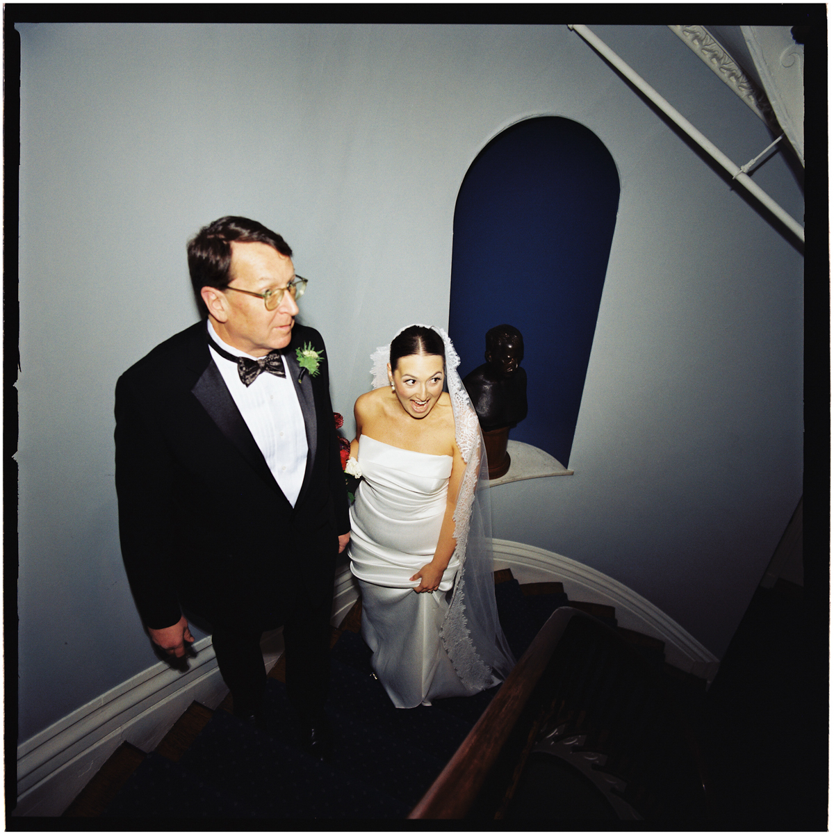 Bride in elegant gown and her father ascending a staircase, with a smile and lively expression, surrounded by decor and an artistic background.