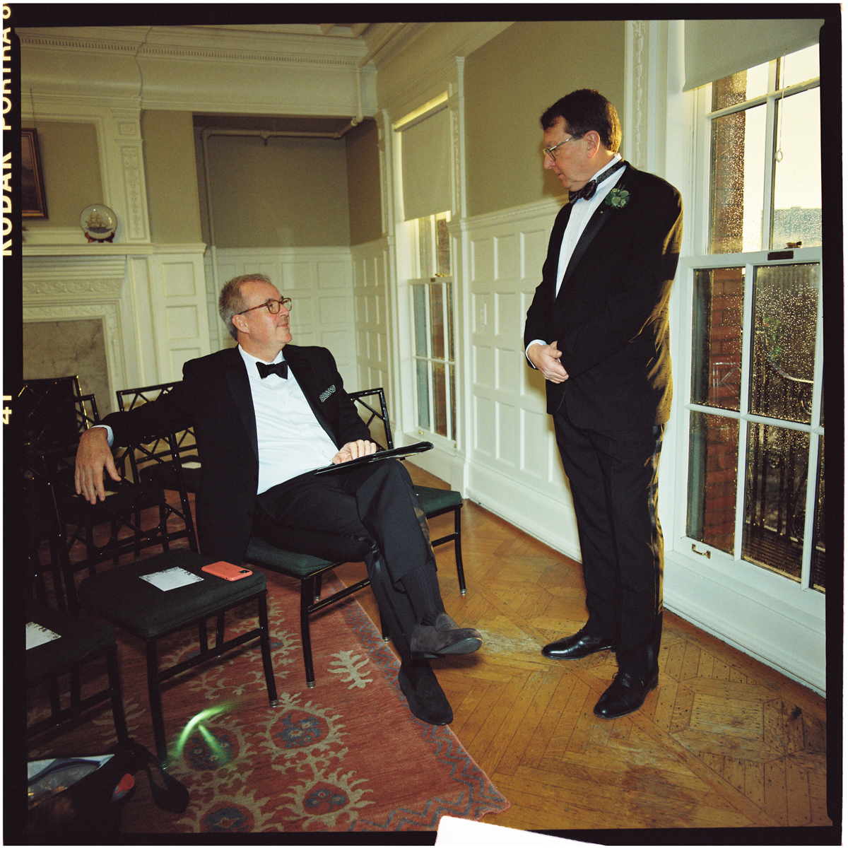 Two men in formal attire converse indoors, one seated and the other standing, with elegant decor and large windows in the background.