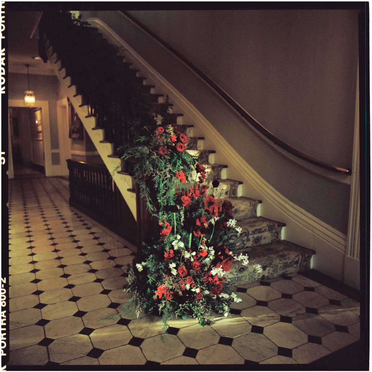 A beautifully arranged display of vibrant flowers at the base of a staircase, featuring red roses and greenery, set in a warmly lit hallway with patterned flooring.