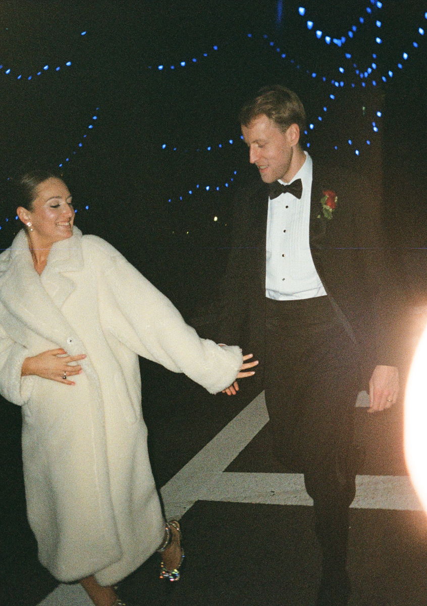 A couple dressed in formal attire enjoying a joyful moment while walking under blue lights in a nighttime setting.