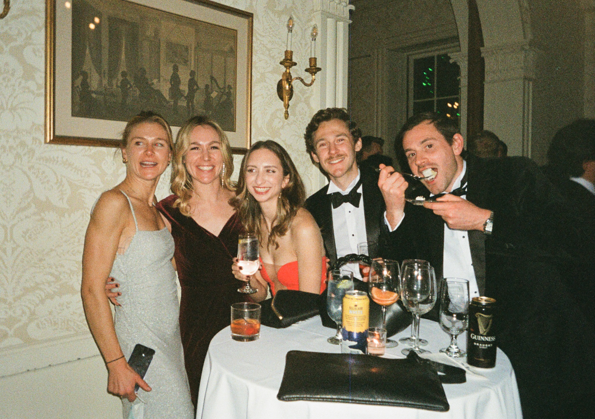 Group of five friends smiling at a table with drinks, dressed in formal attire, enjoying a lively gathering.
