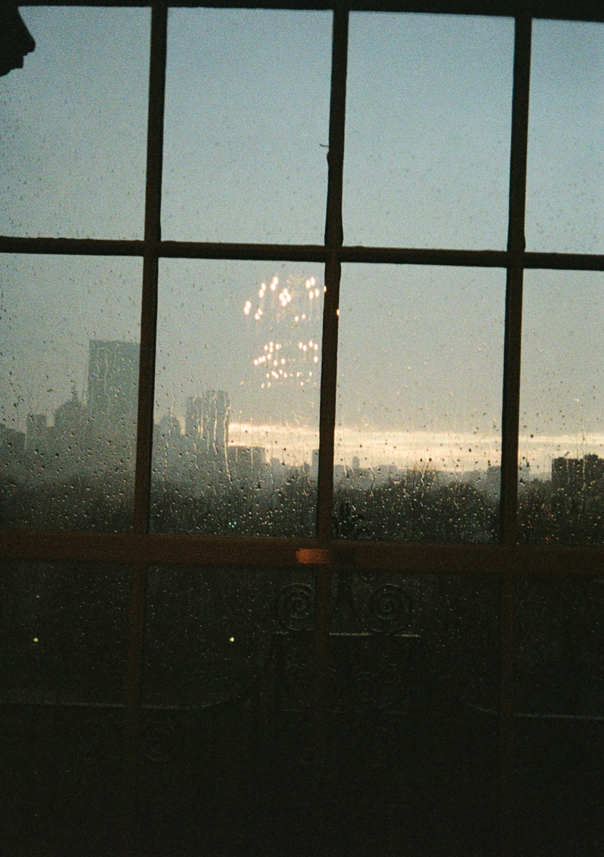 A rainy window reflecting a chandelier, with city silhouettes and a sunset in the background.